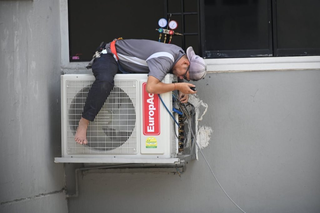 man-in-gray-t-shirt-fixing-the-aircon-7347538 A technician skillfully repairing an outdoor air conditioning unit mounted on a building wall.